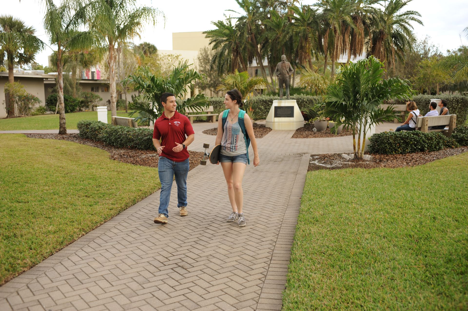 Two students walking and talking on a paved pathway in a tropical campus setting with a statue and seating area in the background